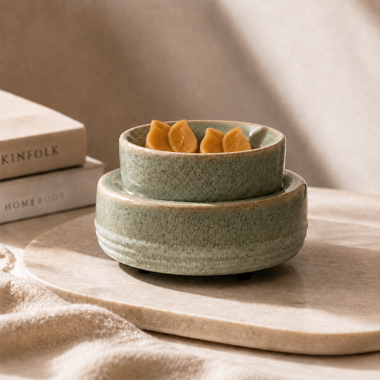 Two stacked speckled sage green ceramic bowls, with the top bowl holding three small orange flower-shaped objects, styled on a light stone tray with neutral fabric and books in the background.