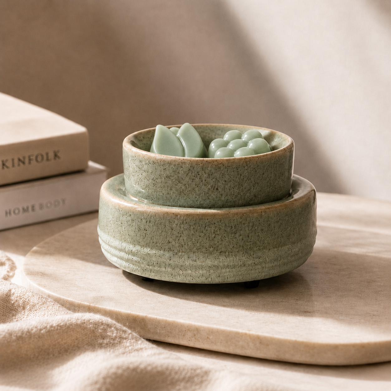 A pair of stacked ceramic bowls in a speckled sage green glaze, with the top bowl containing a small green decorative object, styled on a light stone tray with books in the background.