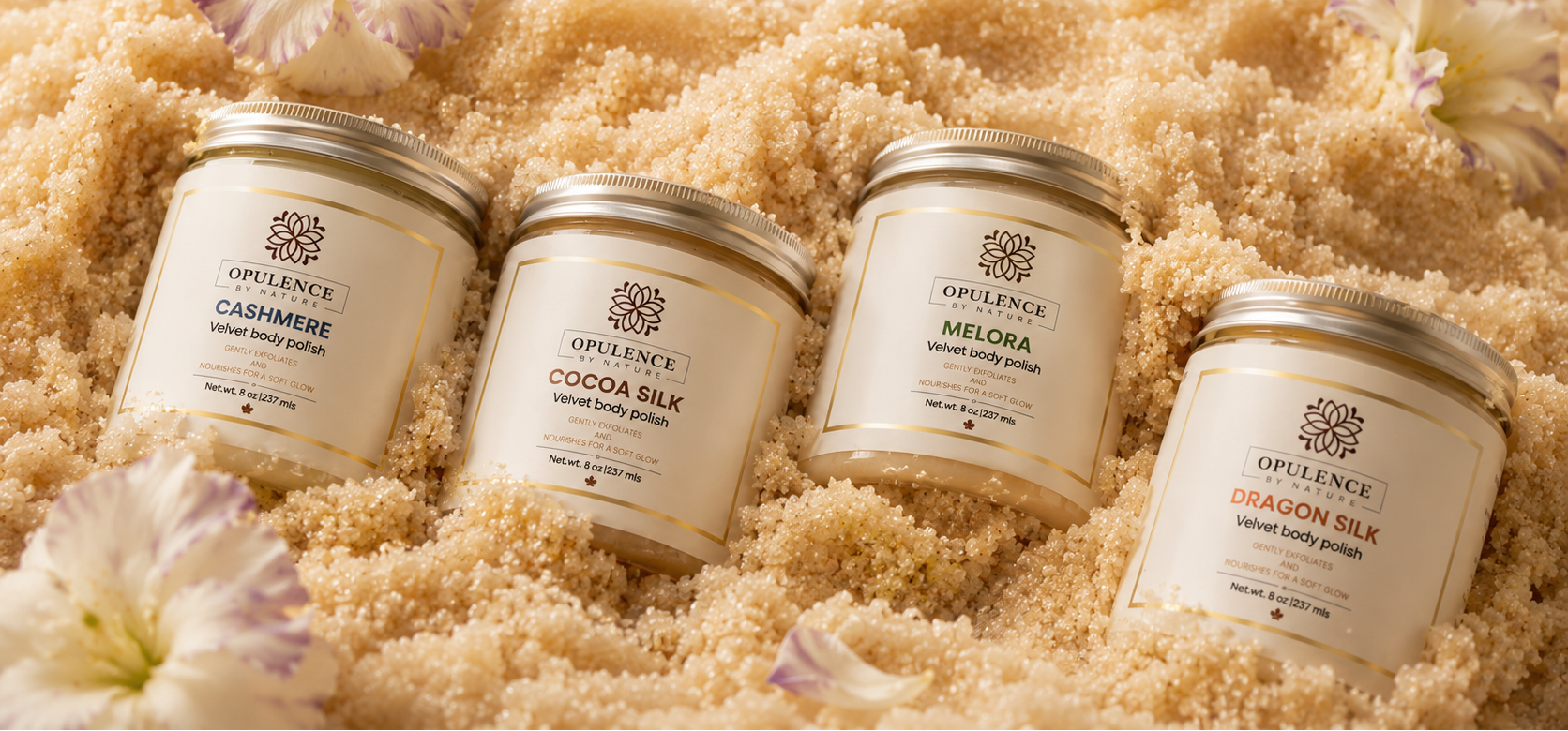 Four Opulence body butter jars arranged on a bed of beige body scrub or sand-like texture with decorative flowers. The jars are cream-colored with gold lids and labels showing different scents: Cashmere, Cocoa Silk, Melora, and Dragon Silk.