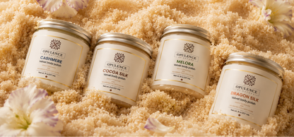 Four Opulence body butter jars arranged on a bed of beige body scrub or sand-like texture with decorative flowers. The jars are cream-colored with gold lids and labels showing different scents: Cashmere, Cocoa Silk, Melora, and Dragon Silk.