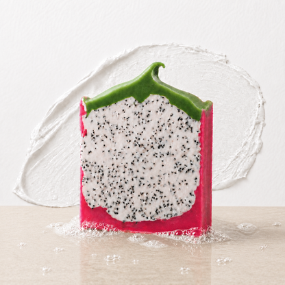 A novelty soap bar shaped like a slice of dragon fruit, featuring a bright pink outer skin, a white flesh section speckled with black seeds, and a green rind, standing on a light beige surface against a white textured background.