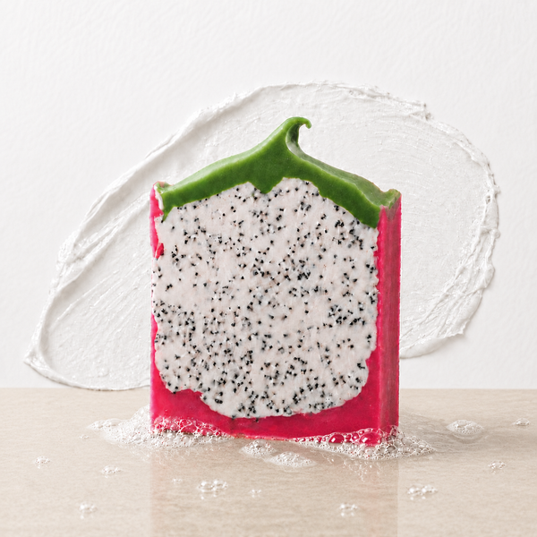 A novelty soap bar shaped like a slice of dragon fruit, featuring a bright pink outer skin, a white flesh section speckled with black seeds, and a green rind, standing on a light beige surface against a white textured background.