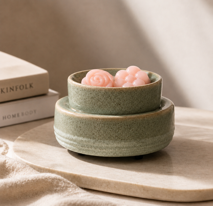 A small speckled ceramic dish in a muted sage green color, shown with a matching shallow bowl on top holding two pink rose-shaped objects, styled on a light stone surface with books in the background.