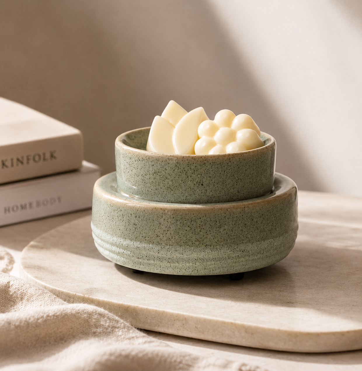 A small speckled sage green ceramic bowl with a matching saucer, styled on a light stone tray with neutral fabric and books in the background. The bowl is filled with cream-colored decorative wax melts or soap pieces.