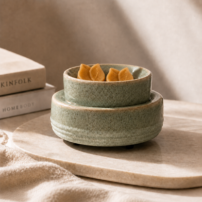 Two stacked speckled sage green ceramic bowls, with the top bowl holding three small orange flower-shaped objects, styled on a light stone tray with neutral fabric and books in the background.