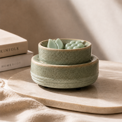 A pair of stacked ceramic bowls in a speckled sage green glaze, with the top bowl containing a small green decorative object, styled on a light stone tray with books in the background.