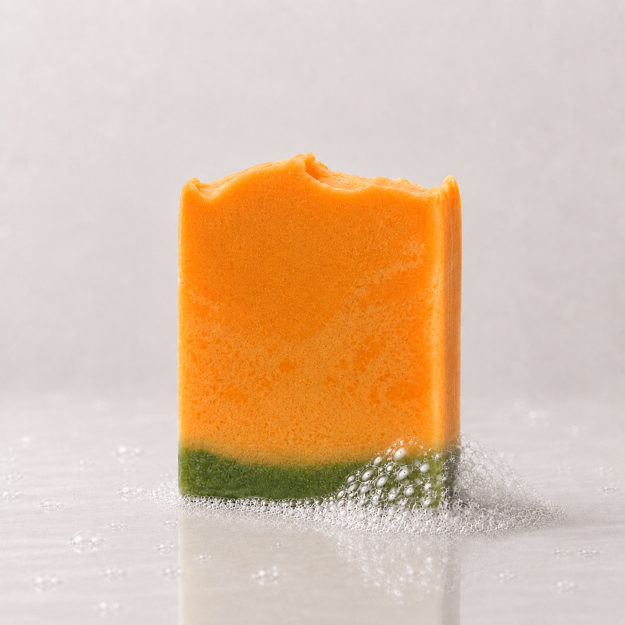 A rectangular bar of soap, featuring a bright orange upper half and a green lower half, standing on a light gray reflective surface with a few bubbles around it.