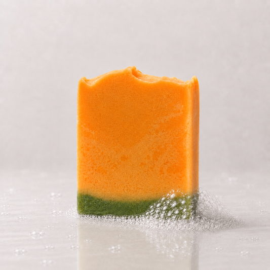 A rectangular bar of soap, featuring a bright orange upper half and a green lower half, standing on a light gray reflective surface with a few bubbles around it.